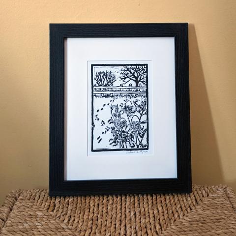 Framed print of a garden in winter, with dried flowers and animal tracks in the foreground, and a fence and some bare trees in the background